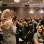 Woman in headscarf addressing crowd sitting at tables