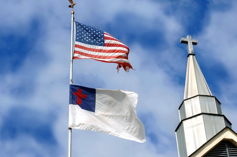 A Christian flag flies beneath the American flag next to a church steeple. nameinfame/iStock via Getty Images Plus
