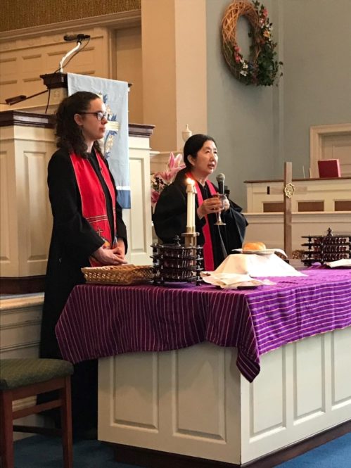 Two female pastors, Kathryn Ray and Yuki Scroggins, stand in front of an altar with bread and grape juice in preparation for Communion.