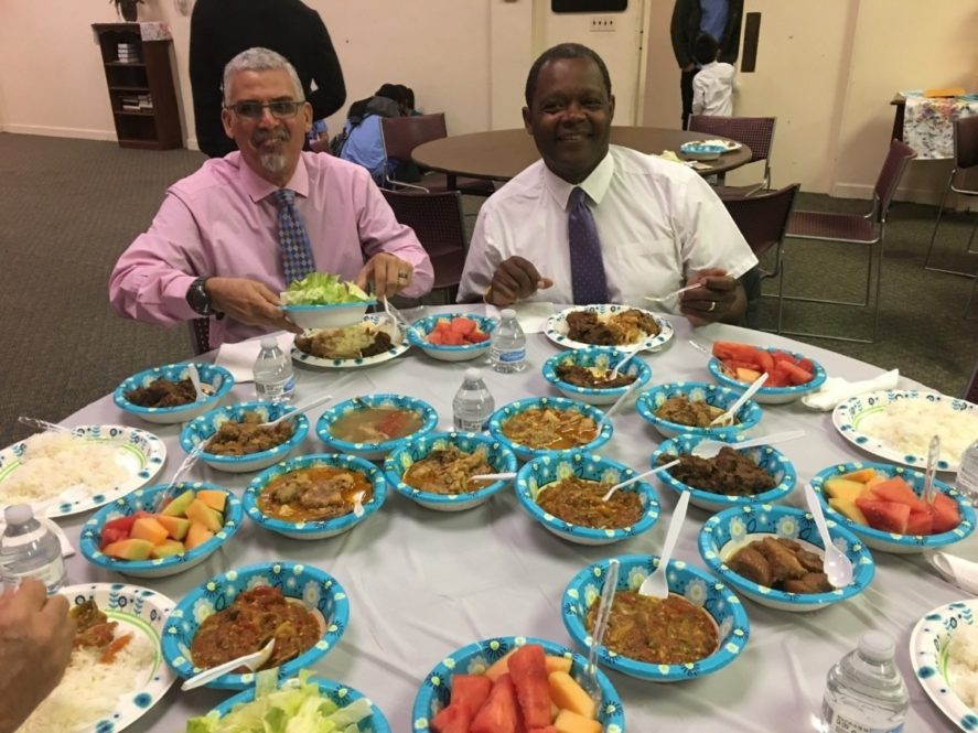 Two male pastors, Juan Angel Gutierrez and Michael Ware smile sitting down to a table full of food. Images provided by Becca Hartman-Pickerill.