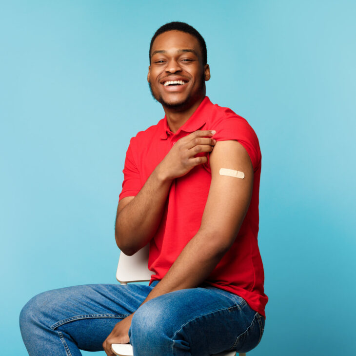 Covid-19 Vaccination. Vaccinated Black Man Showing Arm After Coronavirus Antiviral Injection Sitting With Rolled Up Sleeve On Blue Studio Background, Smiling To Camera