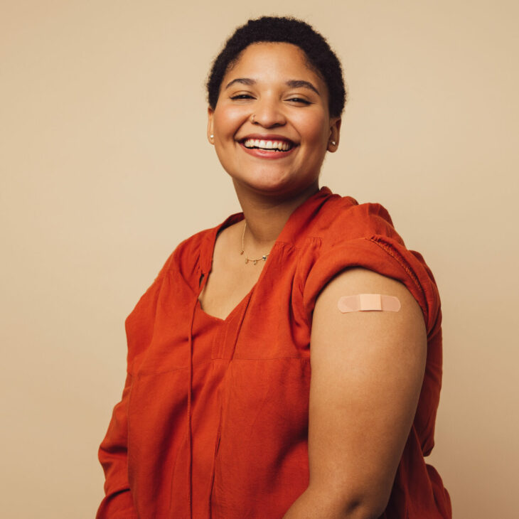 Woman looking happy after getting vaccine shot on brown background. African woman with bandage on her arm after receiving vaccination.