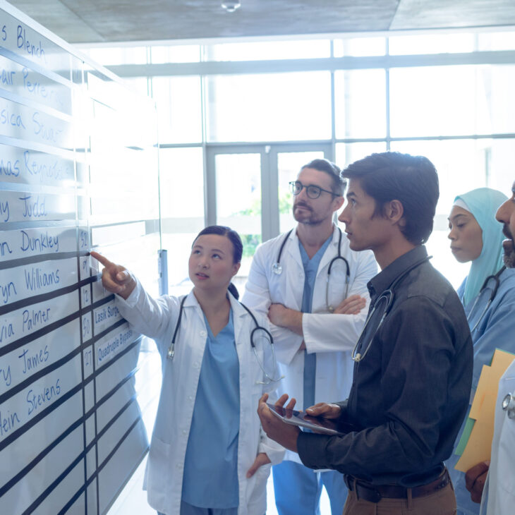Side view of diverse medical team of doctors discussing their shifts on chart at hospital. Asian female surgeon is pointing at the whiteboard.