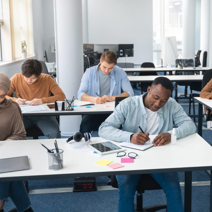 Portrait of diverse group of students sitting at desks in classroom at university, taking test, entrance examination or writing notes in notebook, selective focus