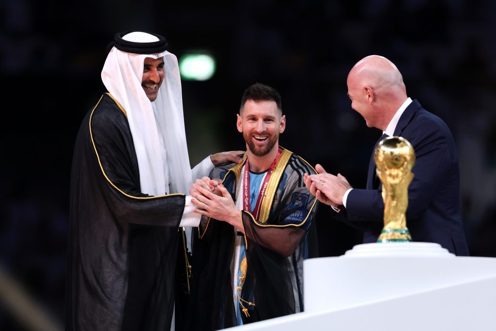 LUSAIL CITY, QATAR - DECEMBER 18: Lionel Messi of Argentina is presented with a traditional robe by Sheikh Tamim bin Hamad Al Thani, Emir of Qatar, while Gianni Infantino, President of FIFA, looks on during the awards ceremony after the FIFA World Cup Qatar 2022 Final match between Argentina and France at Lusail Stadium on December 18, 2022 in Lusail City, Qatar. (Photo by Clive Brunskill/Getty Images)