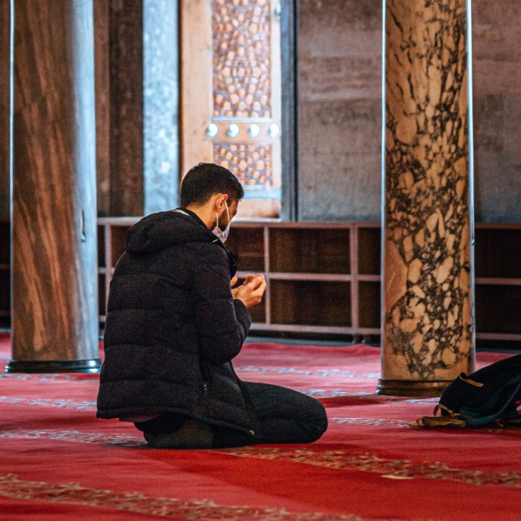 A young muslim man reciting making duaa after prayer while in front of Mihrab of the mosque in the afternoon.