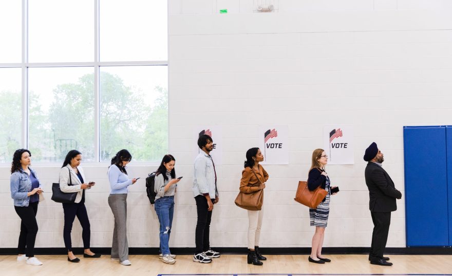 A multiracial group of people, many with smart phones, line up along a wall in a school gymnasium in order to vote.