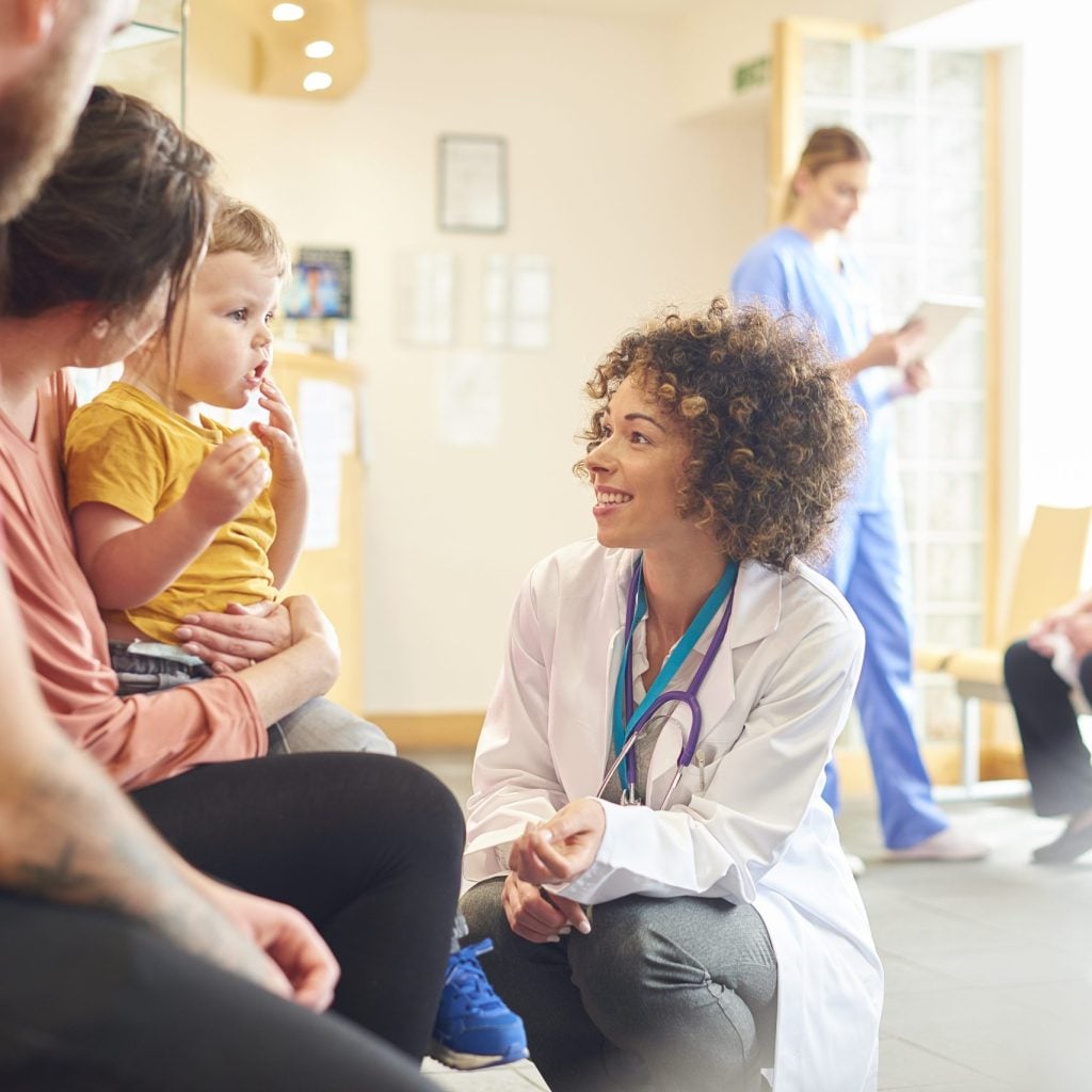 A young family sits talking to the doctor. The toddler is sat on his mother’s knee as the doctor kneels down to talk to him