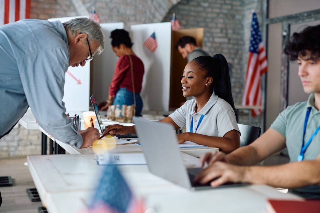Senior US citizen signing up for voting with help of African American female volunteer at polling place.