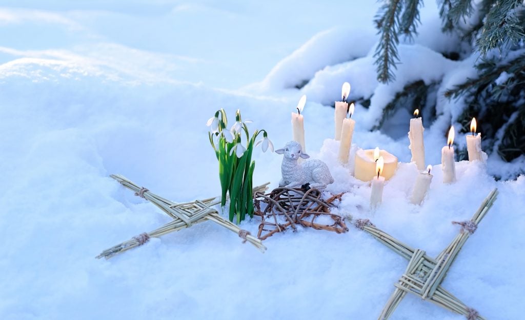 Wiccan altar for Imbolc sabbat. pagan magic ritual. Brigid's cross of straw, candles, snowdrops, toy sheep on snow, winter forest natural background. symbol of Imbolc holiday, spring equinox. (Shutterstock / ju_see)