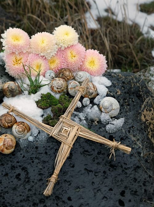 Wiccan altar for Imbolc holiday. Brigid's cross of straw, candles and snail shells on big stone outdoor, abstract nature background. symbol of Imbolc, spring equinox. witchcraft, pagan magic ritual (Shutterstock)